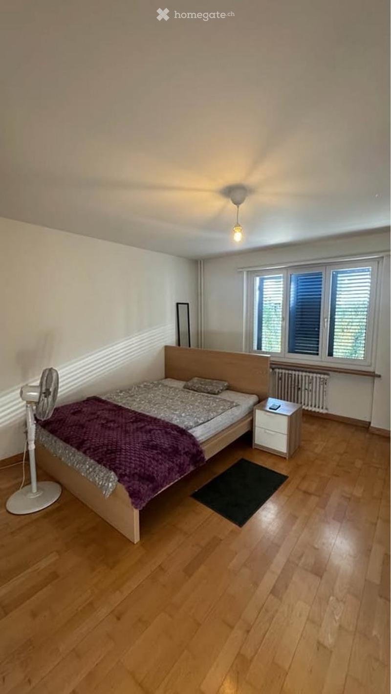 Cozy bedroom with wooden flooring, a bed with a purple blanket, a fan, a nightstand, and a mirror near the window.