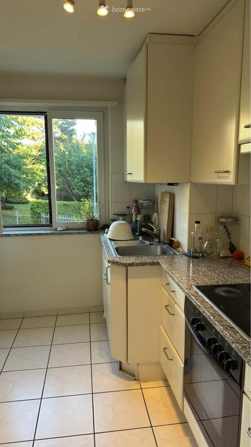 Bright kitchen with white cabinets, granite countertops, tiled floor, and a large window offering a view of greenery outside. Includes a sink, oven, and various small appliances.