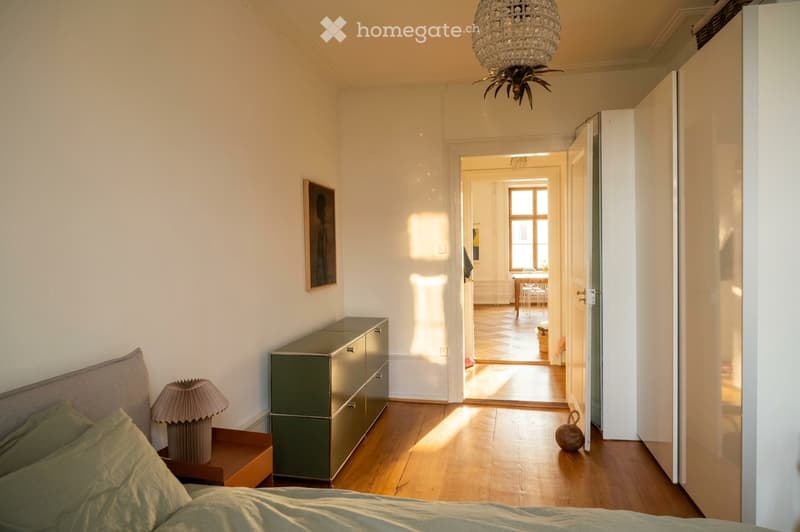 Bright bedroom with wooden flooring, a bed with green bedding, a nightstand with a lamp, a green drawer unit, and an open doorway leading to another room with a table and chairs.