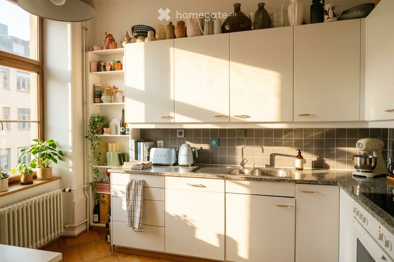 Bright, modern kitchen with white cabinets, granite countertops, stainless steel sink, and various appliances including a toaster and mixer. Shelves hold decorative items and plants, with natural light streaming in from a large window.