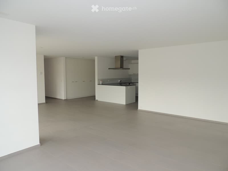 Empty room with light grey tiled flooring, white walls, and a small kitchen area visible in the background.