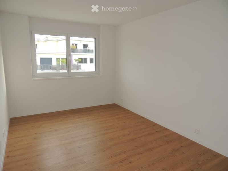 Empty room with wooden flooring and white walls, featuring a large window with a view of a neighboring building and balcony.