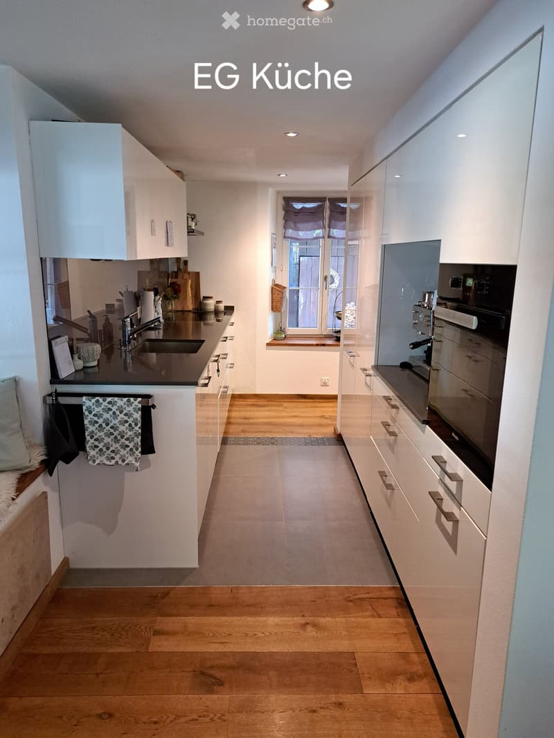 Modern kitchen with white cabinets, wooden flooring, and stainless steel appliances. Features a sink, oven, and various kitchen items on the counter.