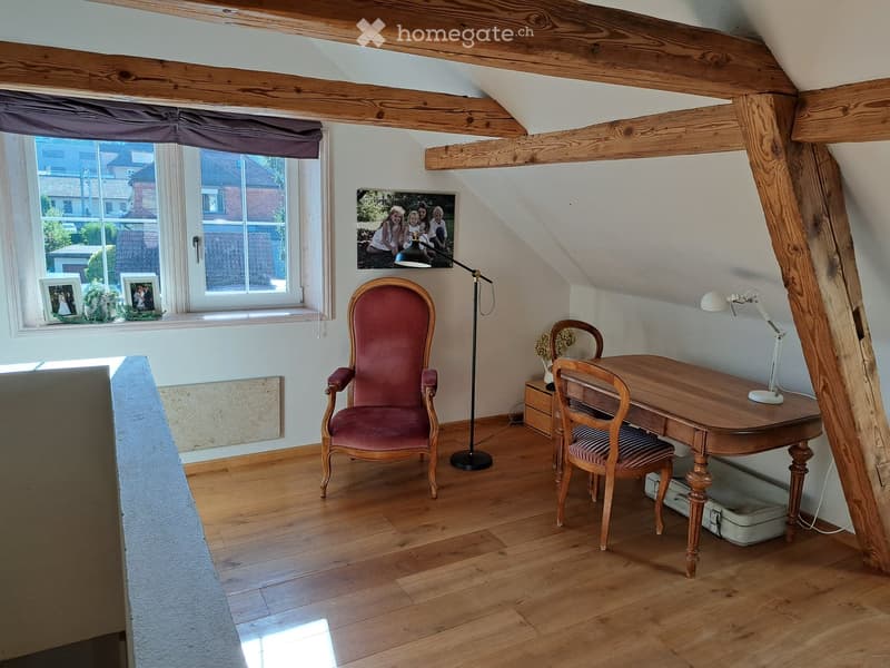 Cozy attic room with wooden beams, hardwood floor, large window, red upholstered chair, wooden desk, and table lamp.