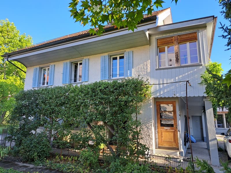 Two-story house with light-colored facade, multiple windows with shutters, wooden front door, and small porch area.