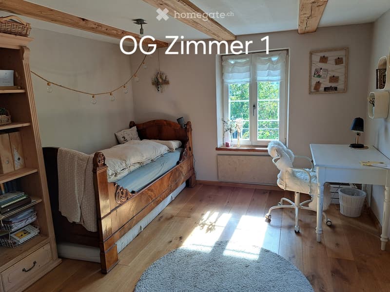Cozy bedroom with wooden bed, desk, chair, bookshelf, and string lights. Features exposed wooden beams and a window with sheer curtains.