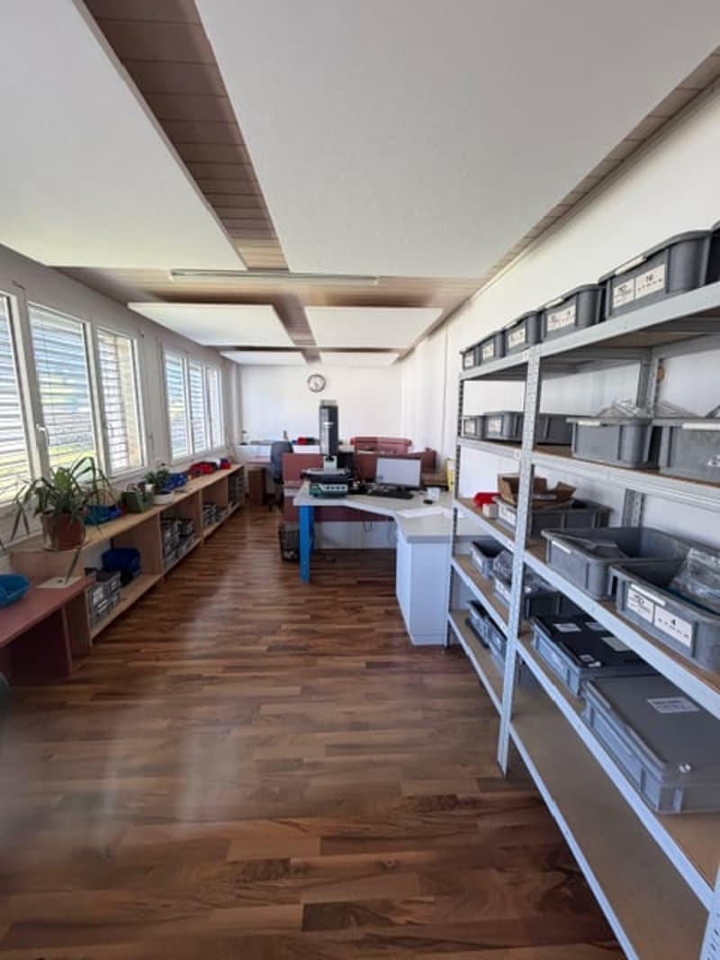 Empty room with wooden floor, white walls, and large windows. There are shelves with storage boxes, a desk with a computer, and a potted plant on a shelf.