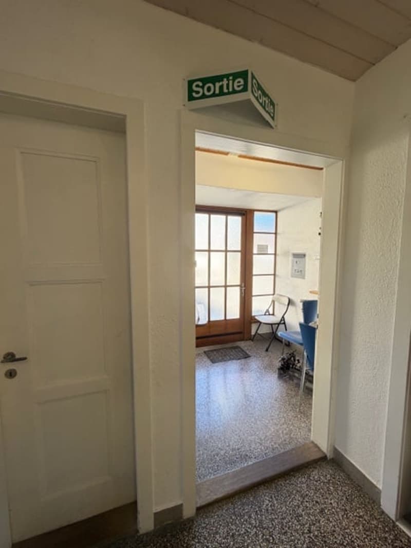 A hallway with a white door, a sign above the door, and a small table with a white cloth. There are two blue chairs and a mat in front of the door.