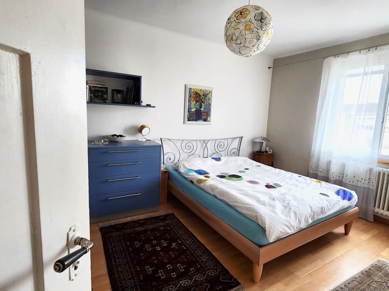 Bedroom with bed, blue dresser, rug, lamp, picture frame, shelf, radiator, window with curtains