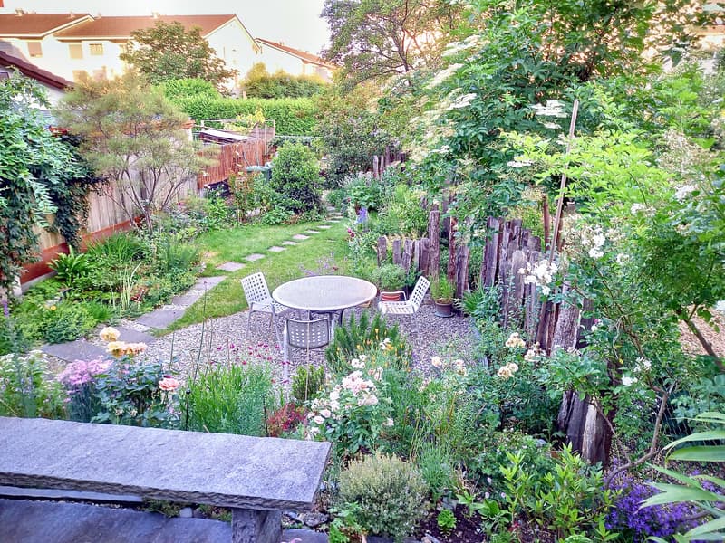 A garden with a table and chairs, surrounded by a variety of plants and flowers, with a gravel path leading to a wooden fence.