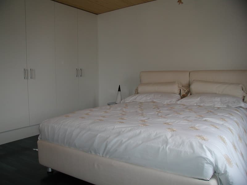 A bedroom with a bed, white walls, white bedspread, white cabinets, nightstand, and a lamp