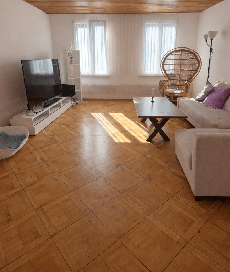 Living room with wooden floor, white walls, large windows, flat screen TV, coffee table, couch, wicker chair, lamp, white shelf, radiator