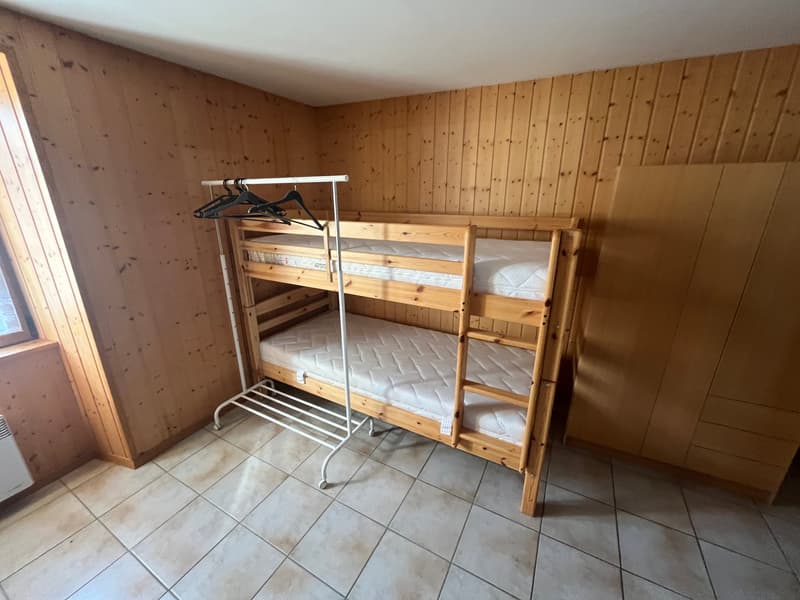 A room with wooden walls, a wooden bunk bed, a white metal clothes rack, a wooden cabinet, and a white tiled floor.