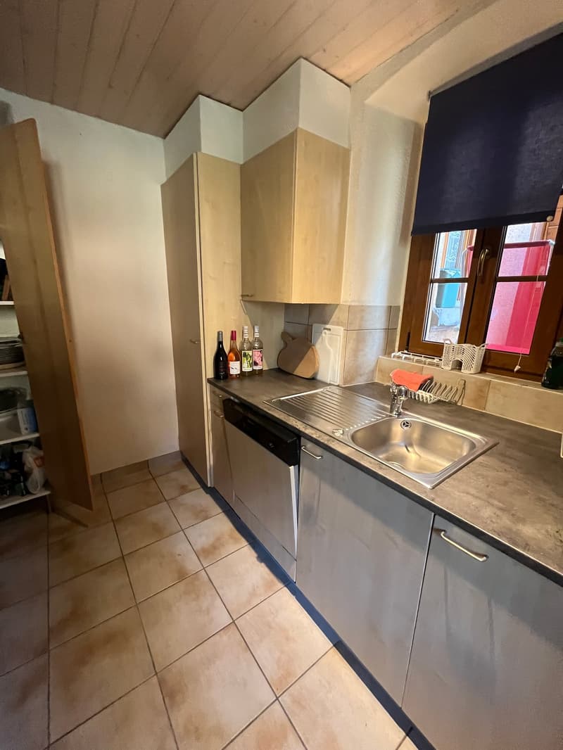 kitchen with tiled floor, stainless steel sink, dishwasher, cabinets, open door to pantry, bottles on counter, window with curtain