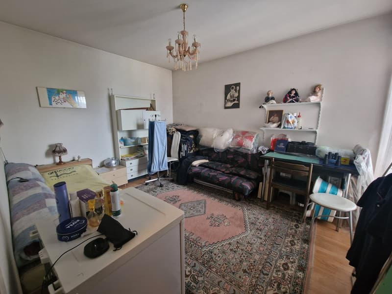 Bedroom with bed, desk, couch, shelves, rug, chandelier, and various items on the desk and floor.