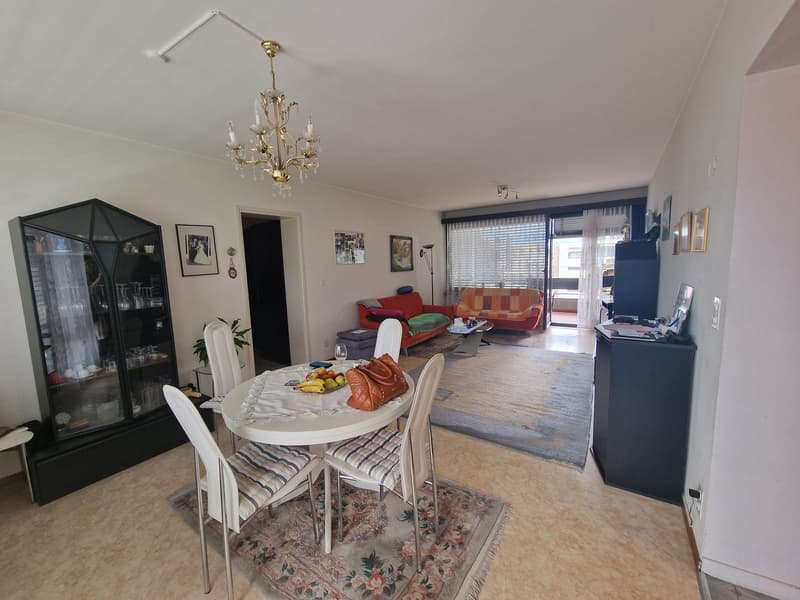 Dining room with a round table, chairs, a chandelier, a cabinet, a rug, a couch, a coffee table, a rug, a plant, a lamp, a TV, and a balcony