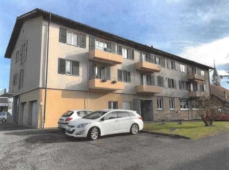 A three-story apartment building with beige and grey walls. There are multiple balconies visible, and two cars are parked in front. A small tree is on the grass, and a bench is beside it.