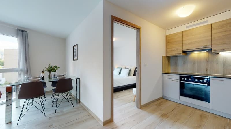 Open floor plan, dining table with chairs, modern kitchen with white cabinets and stainless steel stove, white and wooden flooring, large window with curtain, bedroom with bed and pillows