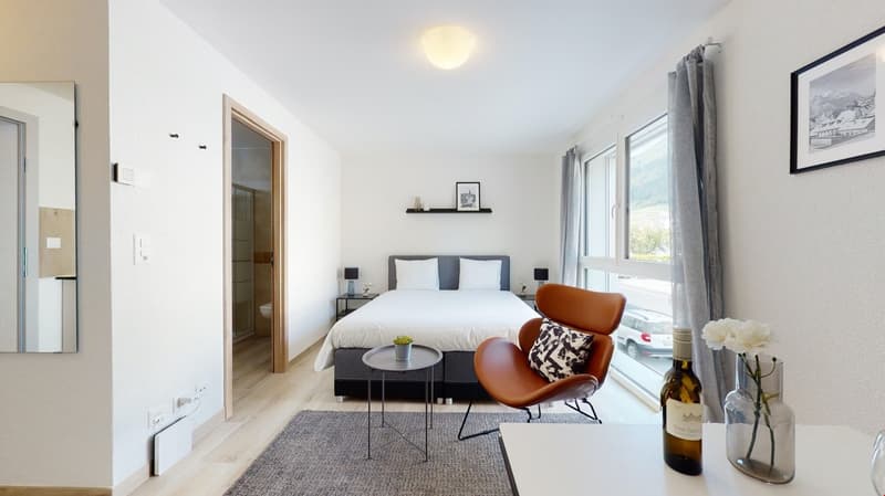 Bedroom with a bed, chair, nightstands, a carpet, a table with a flower vase and wine bottle, and a large window