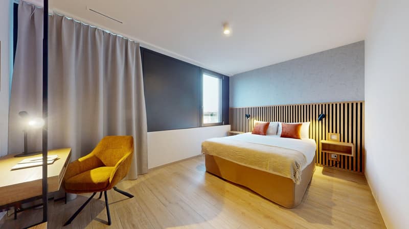 Spacious bedroom with wooden flooring, gray and blue color scheme, large window, yellow accent chair, wooden headboard with horizontal slats, bedside tables, and a double bed with yellow and white bedding.