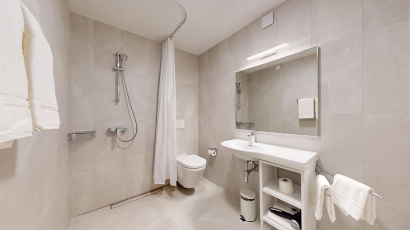 Spacious bathroom with beige tile walls, a large vanity with a sink, a toilet, and a shower with a curtain. The bathroom also has a towel rack and a small storage cabinet.