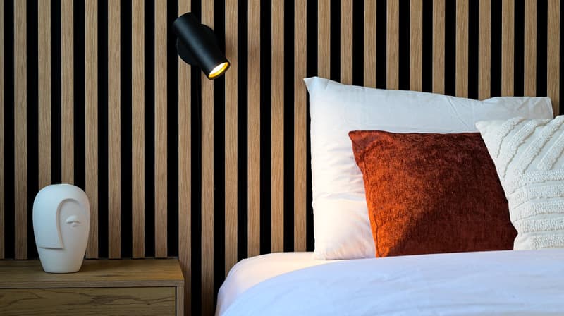 Wooden paneled wall, white bedding, orange throw pillow, bedside table with lamp