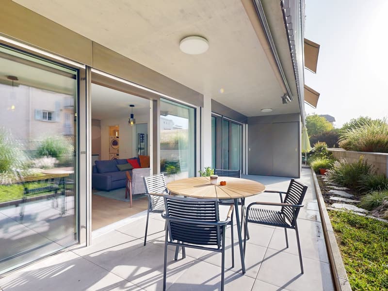 Modern house with glass doors leading to a balcony, featuring a wooden table with chairs, glass windows, and indoor plants, a couch, and a kitchen area