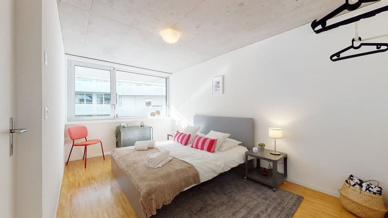 Spacious bedroom with large window, hardwood floor, gray bed frame, red accent chair, side table with lamp, and storage cabinet
