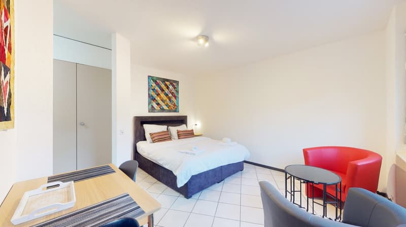 Spacious bedroom with a double bed, colorful artwork on the wall, and modern furniture including a red armchair and a small dining table with stools.