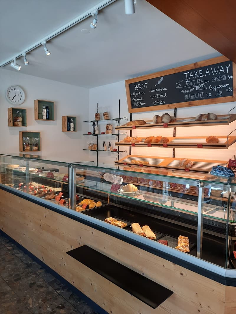 A bakery counter with a glass display case, shelves with bread, a chalkboard sign for TAKEAWAY, wooden display cases, and framed clocks