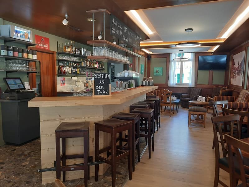 Bar counter with stools, shelves with liquors and glasses, a blackboard menu, digital register, tables and chairs, a television
