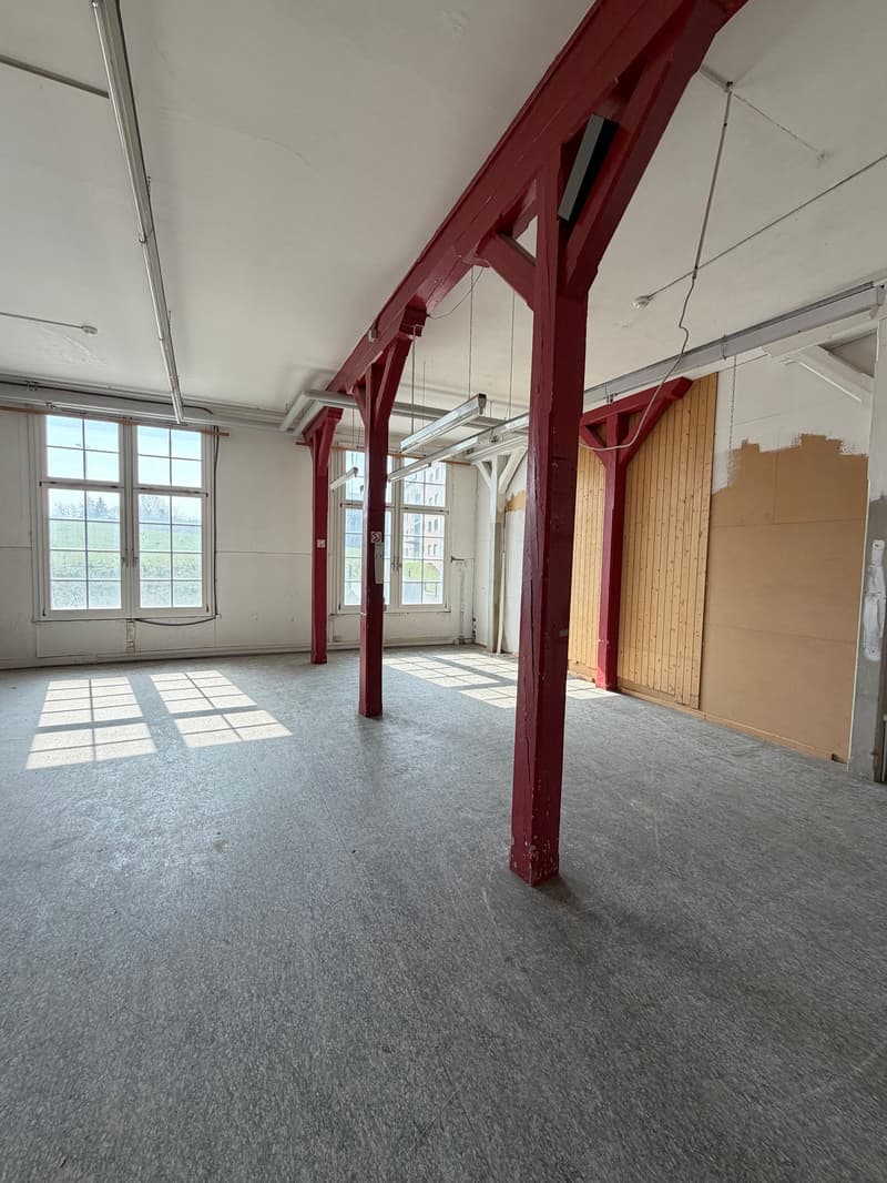 Empty room with red pillars, white ceiling, gray floor, large windows, and some exposed wires