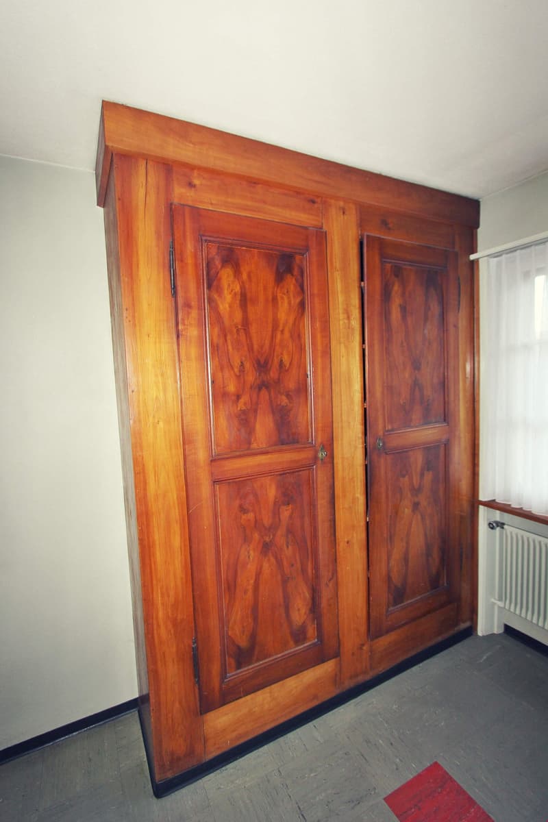 large wooden wardrobe, two doors, wooden floor, white wall, window with white curtains