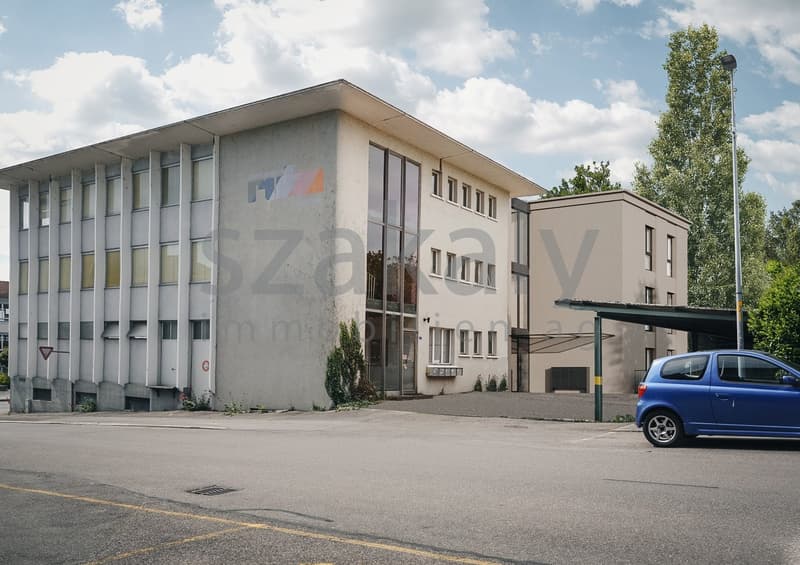A multi-story commercial building with a modern, minimalist design. The building has large windows and a prominent logo on the facade. There is a parking lot in front of the building, and a blue car is visible in the lot.