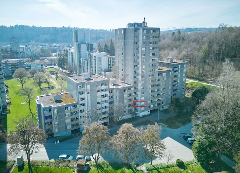 Aerial view of a multi-story residential building with several floors, surrounded by trees and grass, with a parking lot in front.