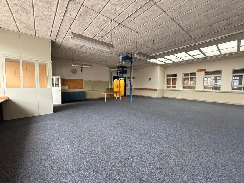 Empty room, carpeted floor, fluorescent ceiling lights, wooden table, white walls, arched windows, wooden cabinets, door.