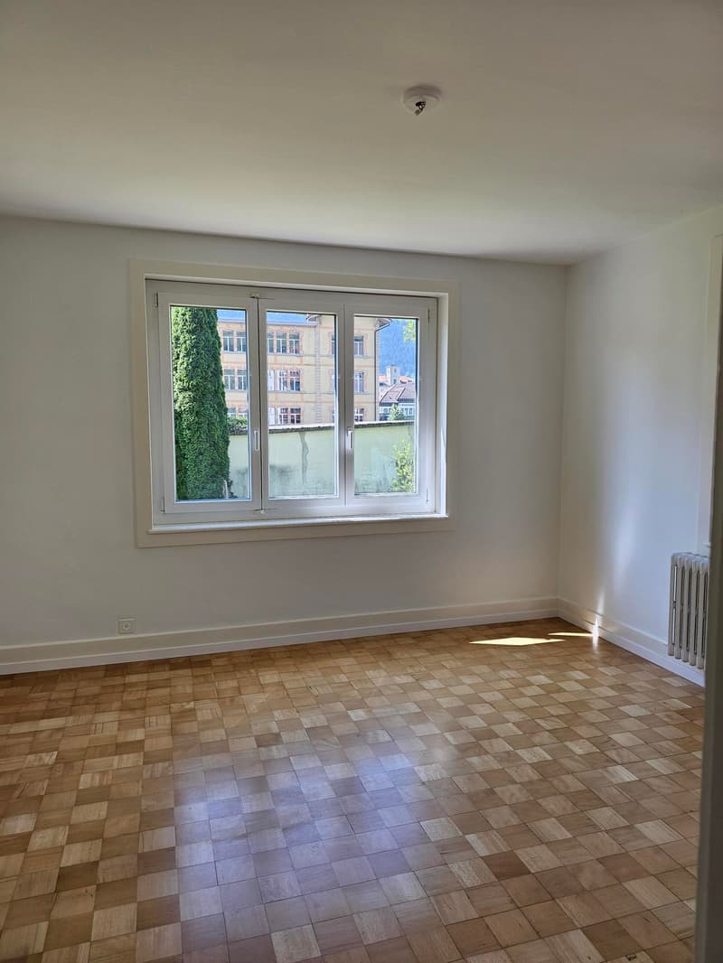 This image shows an empty room with white walls and a large window overlooking a city street. The floor is covered with a patterned tile or wood flooring. The room appears to be in a residential or apartment building setting.