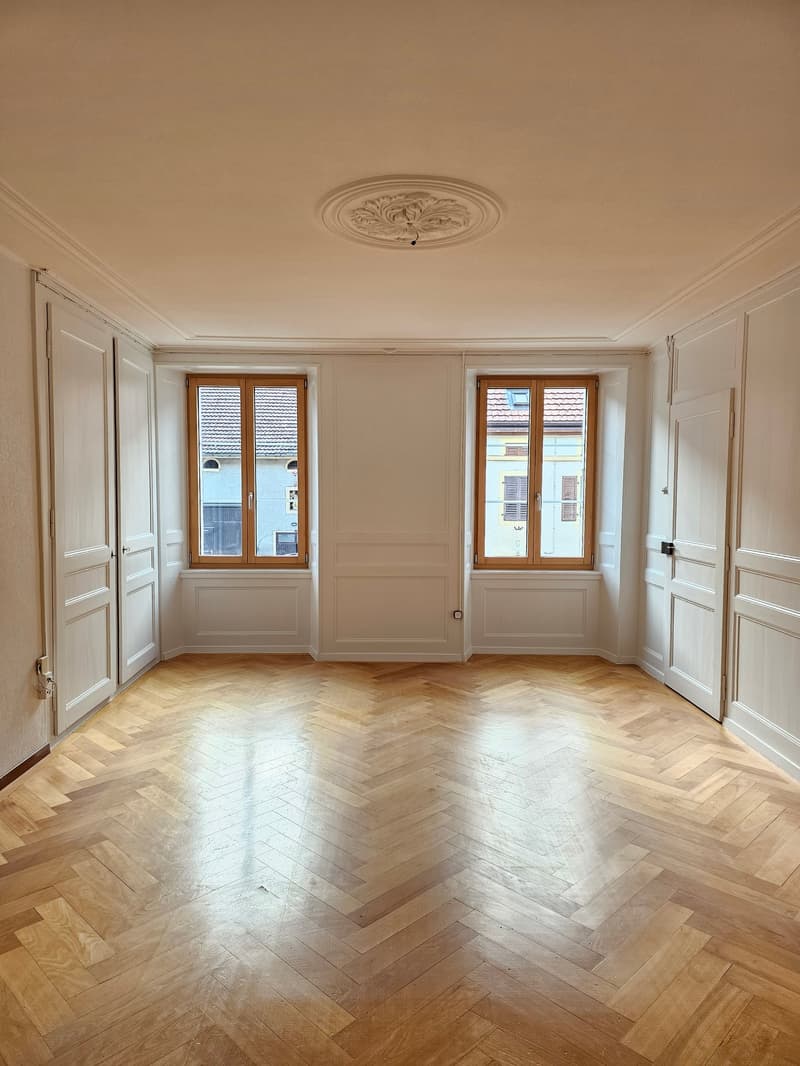 Empty room with wooden herringbone flooring, white walls, 2 windows, ornate ceiling medallion, decorative door frames
