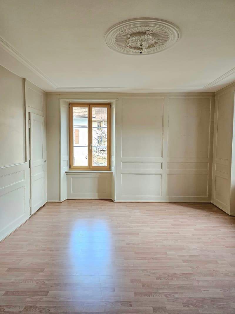 spacious room, white walls, wooden floor, central window, ceiling medallion, doors