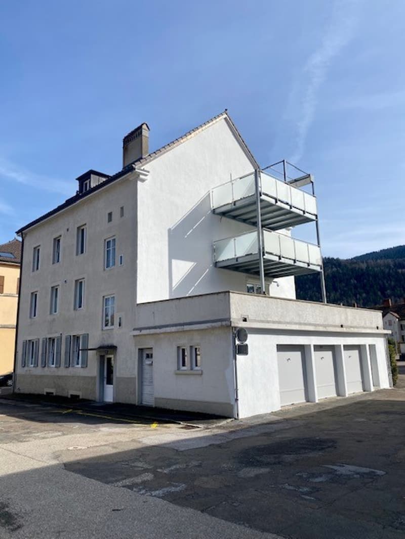 Three-story building with white exterior, balcony on the second floor, garage doors, clear blue sky, and mountain view