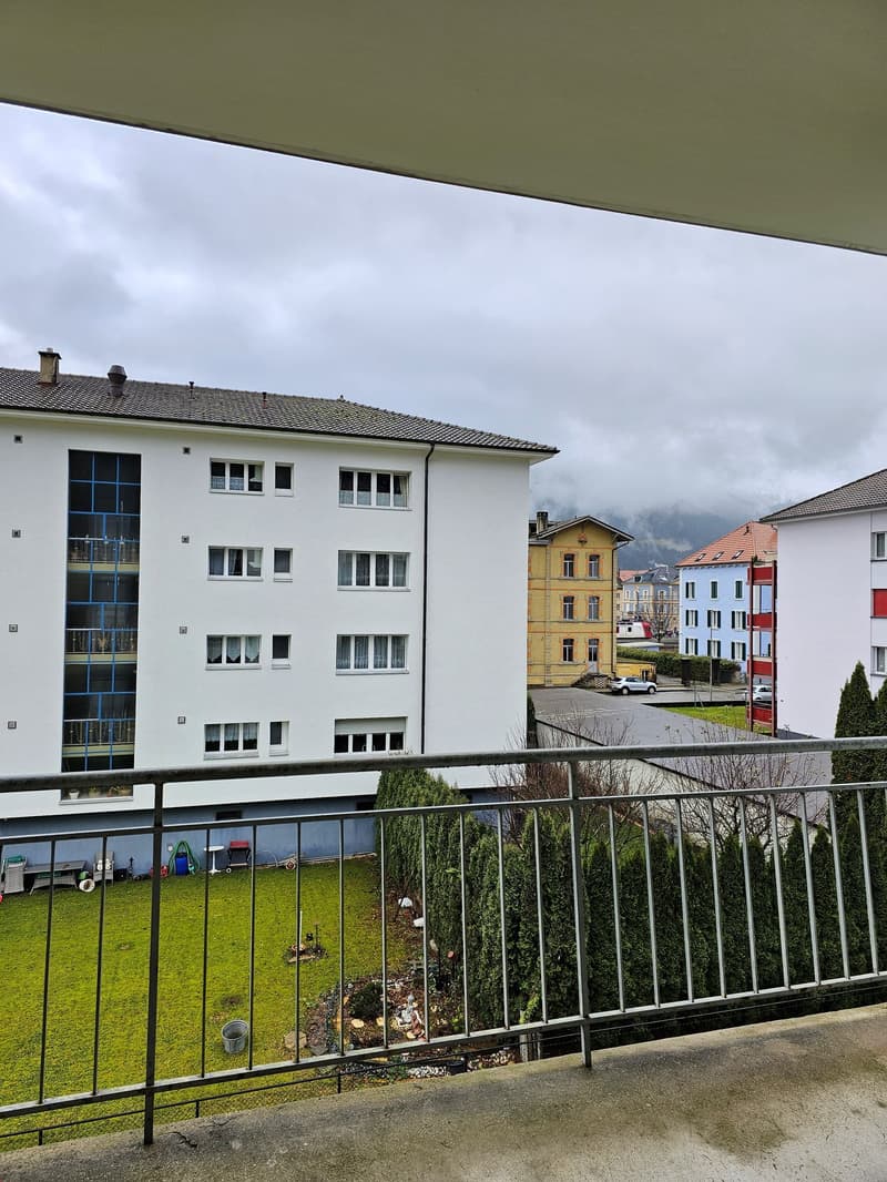 Large balcony, white apartment building, grey parking area, cars, mountains in distance