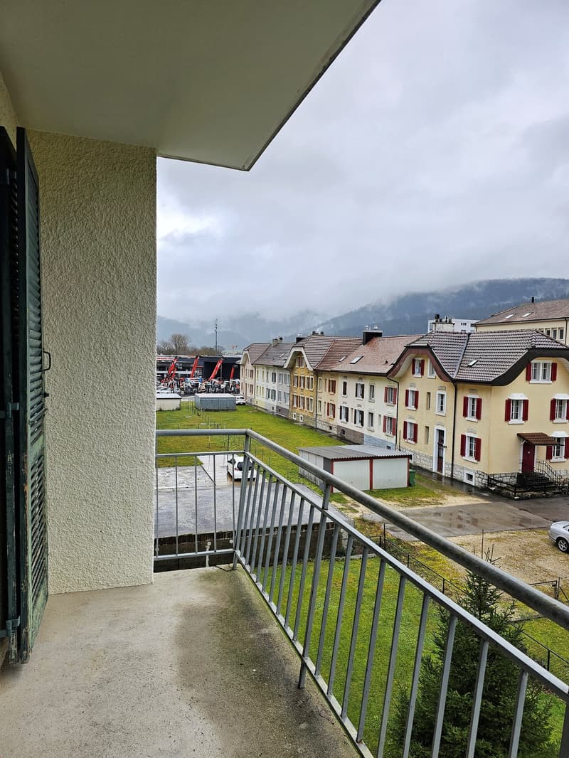 A balcony of a house with railings, overlooking a small lawn, a white car, and other buildings.