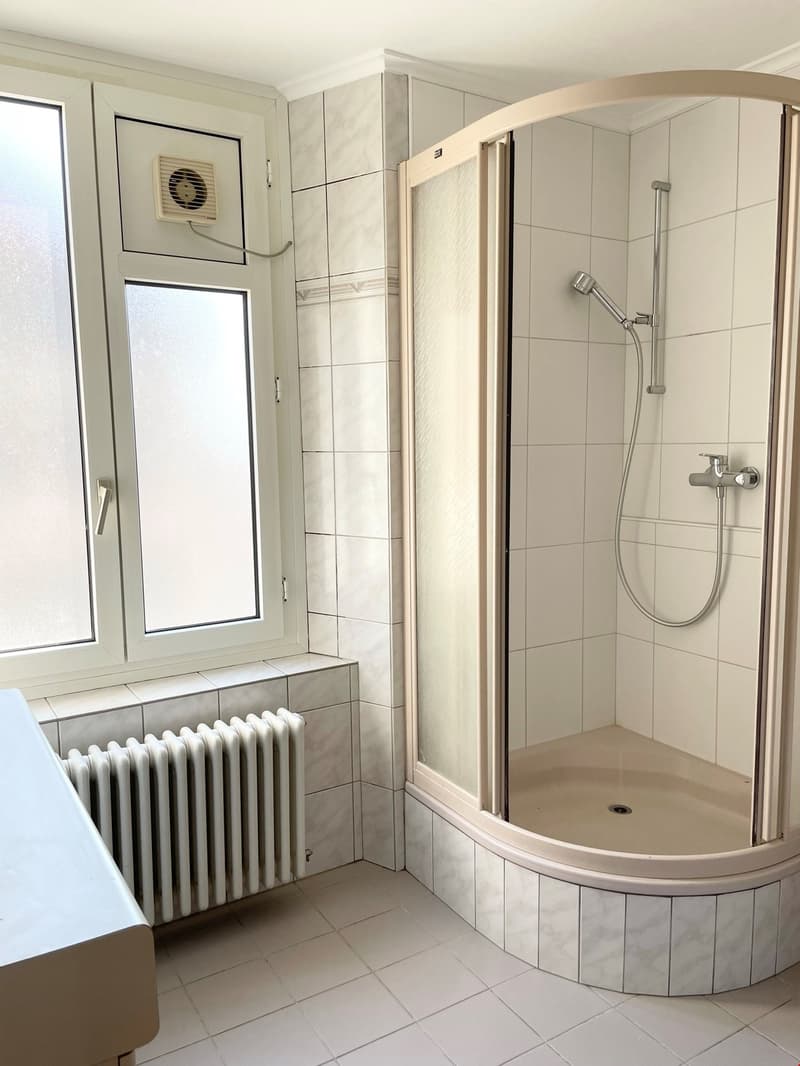 Modern white tiled bathroom with a glass enclosed shower and a white sink, an extractor fan on the wall and a white radiator