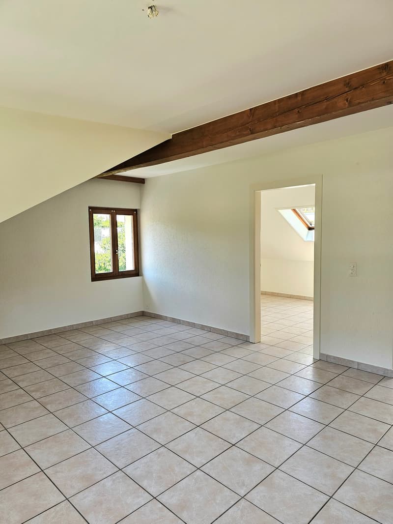 This image shows a spacious room with a slanted ceiling and exposed wooden beams. The room has a tiled floor and a window that provides natural light. The room appears to be empty, with no furniture or decor visible.