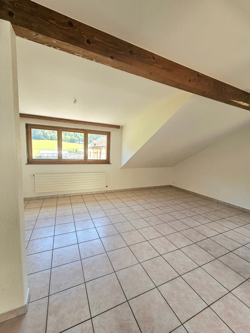 Spacious attic room with exposed wooden beams, large windows providing natural light, and tiled flooring.