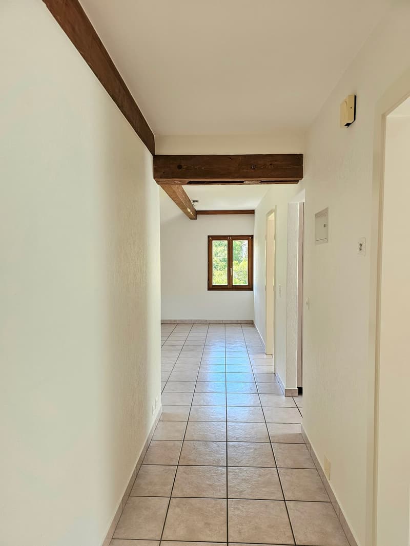 The image shows a hallway with a tiled floor and exposed wooden beams on the ceiling. The walls are painted a light color, and there is a window at the end of the hallway providing natural light. The hallway appears to lead to other rooms or areas of the