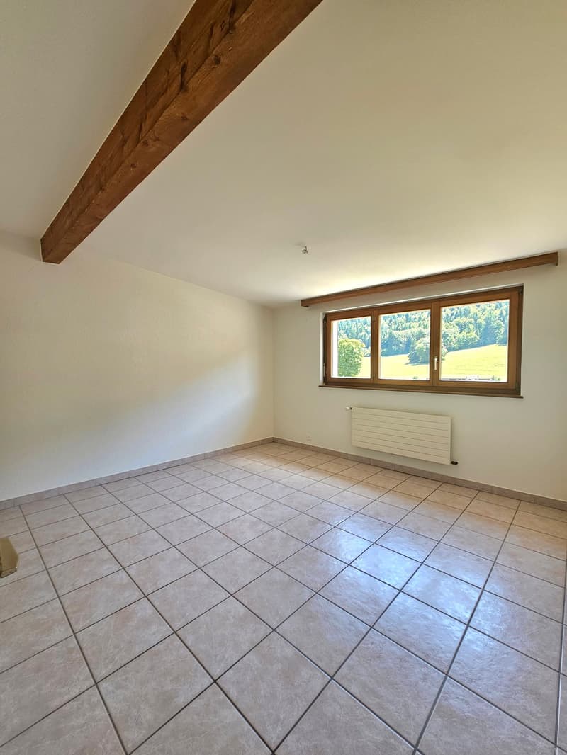 This image shows a spacious room with a tiled floor and a large window overlooking a scenic outdoor view. The room has a wooden beam running across the ceiling, adding a rustic touch to the space. The walls are painted a neutral color, creating a clean an