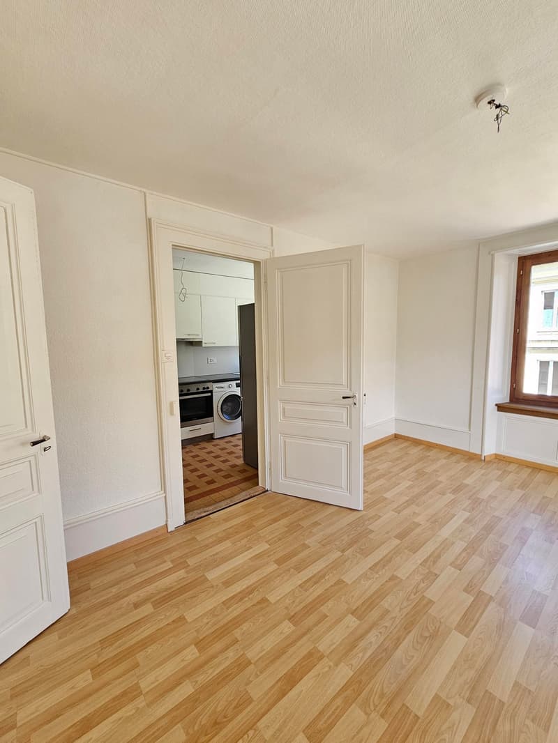 Empty room with wooden floor, white walls, open door, kitchen with washing machine and oven