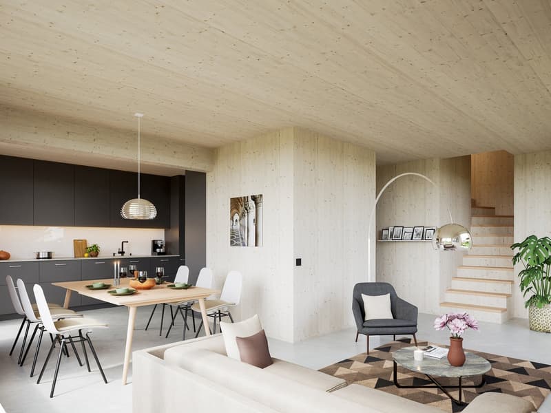 Open-plan living and dining area with wooden table, white chairs, gray armchair, beige sofa, wooden floor, white walls, wooden ceiling, pendant light, coffee table, flowers, staircase, kitchen with black cabinets, countertop, and appliances