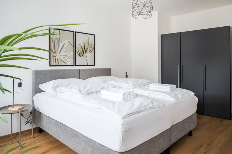 Spacious bedroom with a gray upholstered bed frame, white bedding, and a large black wardrobe. The room features framed artwork on the walls, a geometric pendant light, and a potted plant, creating a modern and minimalist aesthetic.
